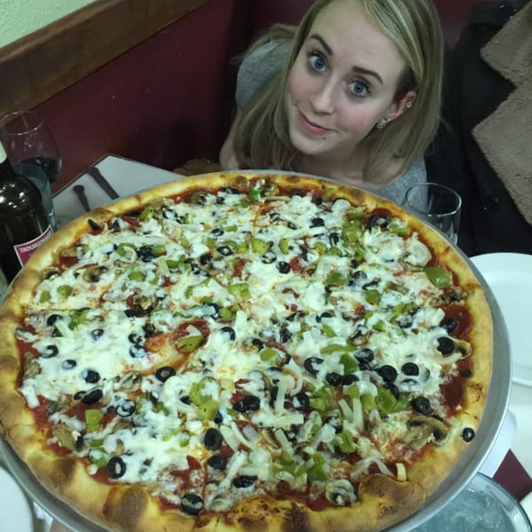 A woman smiles behind a large Lizzano's Pizza with black olives, green peppers, and mushrooms.