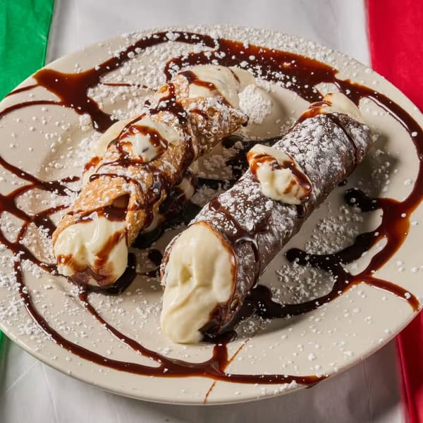 Two Lizzano's Pizza cannolis, one chocolate and one classic, drizzled with syrup and powdered sugar.