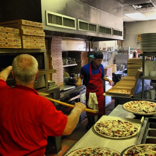 Two Lizzano's Pizza chefs preparing pizzas in a busy kitchen with an oven and pizza boxes.